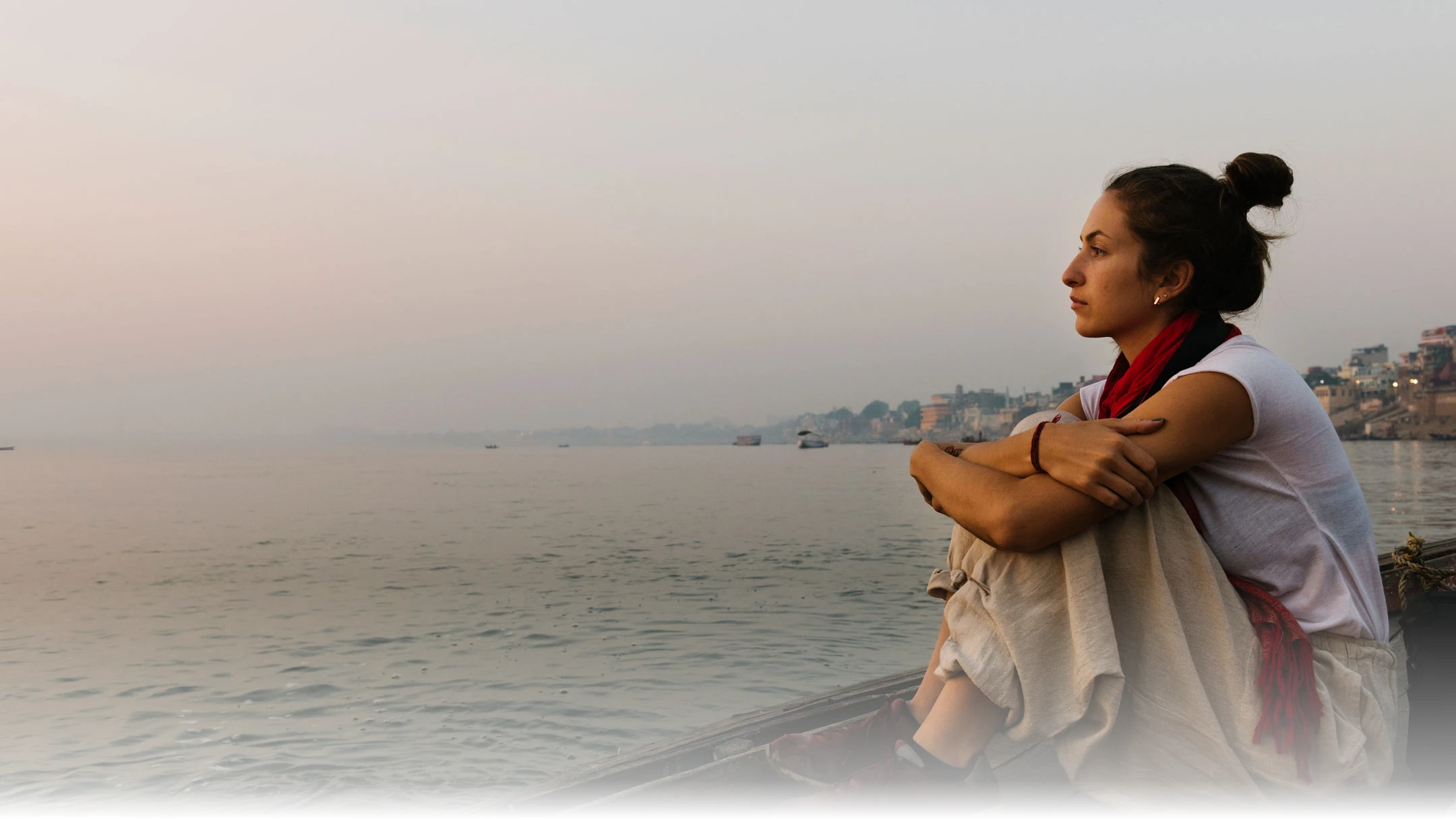 A woman sits alone on a boat at dawn, looking out across a still river toward the ghats